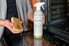 A person holding a bottle of Delphis Heavy Duty Degreaser Spray next to an oven door, with their other hand holding a stained sponge.