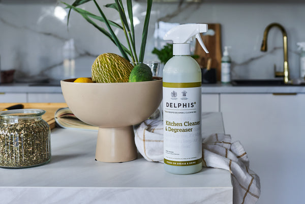 A bottle of Delphis Kitchen Cleaner & Degreaser standing on a white countertop in a modern kitchen setting, next to a beige pedestal bowl with decorative items and a striped cloth.