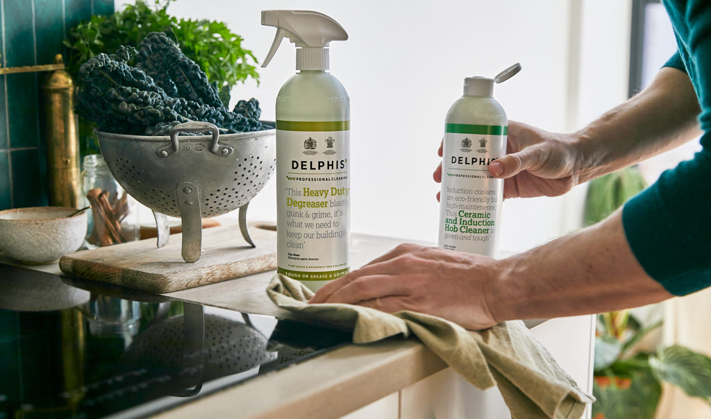 A person in a teal shirt cleans a dark countertop with a cloth while holding a Delphis cleaning spray bottle. A matching Delphis cleaner bottle and a metal colander with leafy greens sit on the counter in a bright kitchen setting.