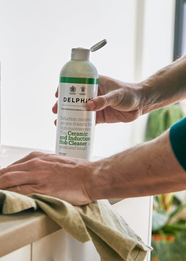 A person in a teal shirt cleans a dark countertop with a cloth while holding a Delphis cleaning spray bottle. A matching Delphis cleaner bottle and a metal colander with leafy greens sit on the counter in a bright kitchen setting.