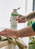 A person in a teal shirt cleans a dark countertop with a cloth while holding a Delphis cleaning spray bottle. A matching Delphis cleaner bottle and a metal colander with leafy greens sit on the counter in a bright kitchen setting.